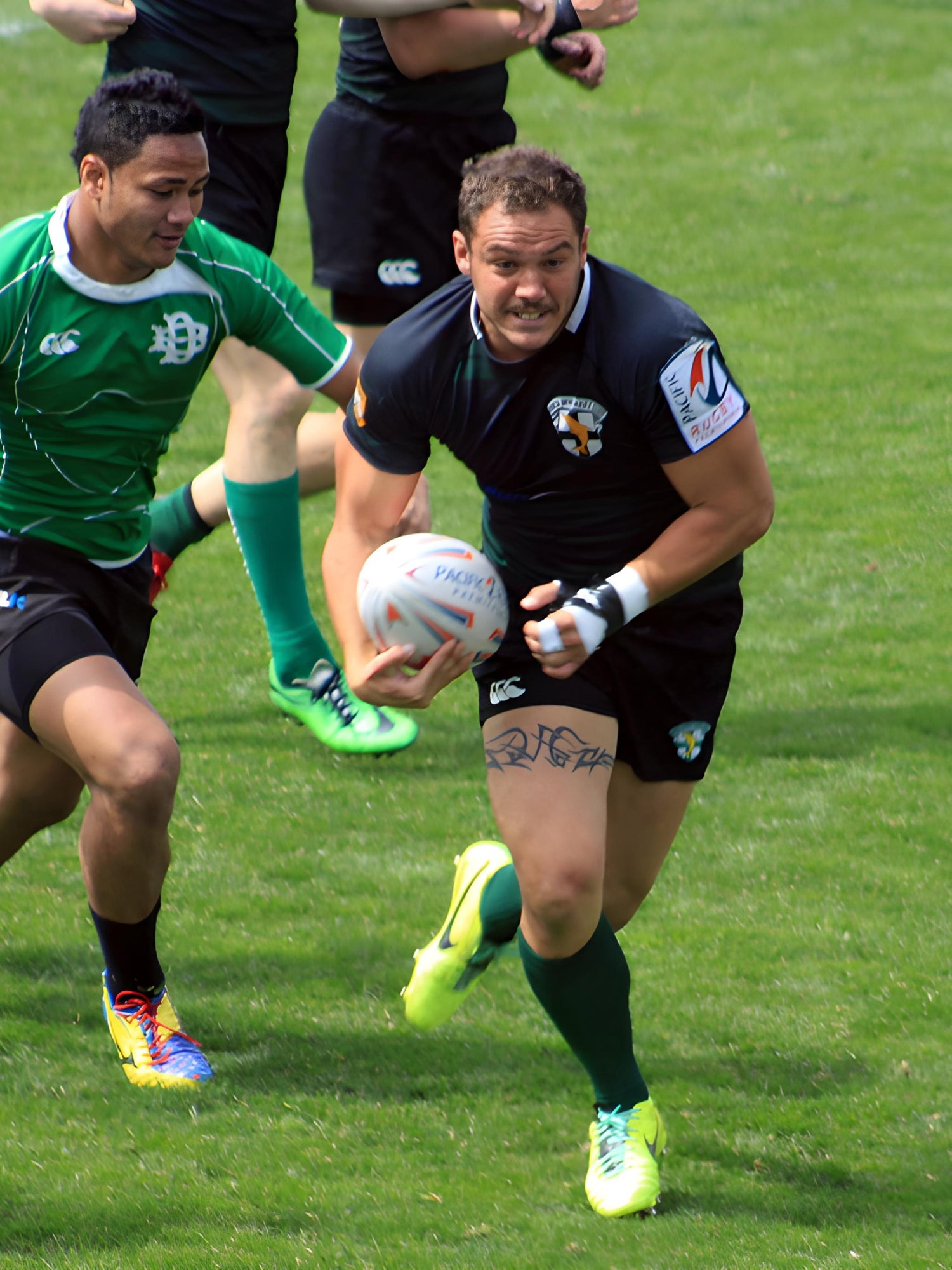 Rugby player, Mathieu Lesgourgues, in black running with the ball on a grass field, pursued by a player in green.